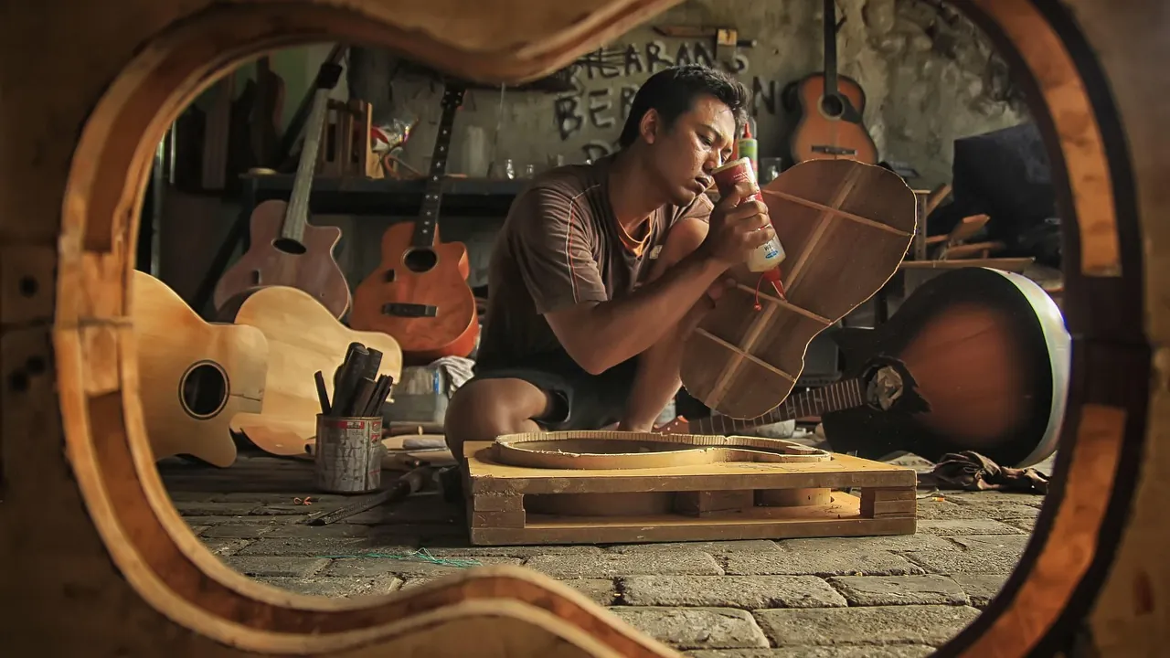 Un luthier ponce une caisse de guitare dans son atelier, vu à travers une caisse inachevée